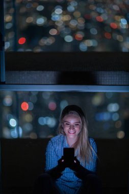 Geceleri arka planda şehir manzarası olan akıllı telefon kullanan genç bir kadın. Yüksek kalite fotoğraf. Cep telefonu, teknoloji, konsept. Yüksek kalite fotoğraf