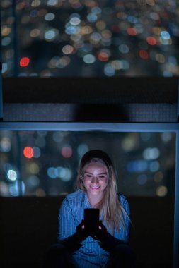 Geceleri arka planda şehir manzarası olan akıllı telefon kullanan genç bir kadın. Yüksek kalite fotoğraf. Cep telefonu, teknoloji, konsept. Yüksek kalite fotoğraf