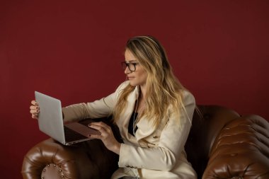 Evde dizüstü bilgisayar ve gazetelerle çalışan genç bir kadın klasik bir kahverengi koltuk ve kulaklıkla çalışıyor. Kırmızı boyalı duvar arka planı. Merkez ofis konsepti. Çalışmak için gri defter. Yüksek kalite fotoğraf