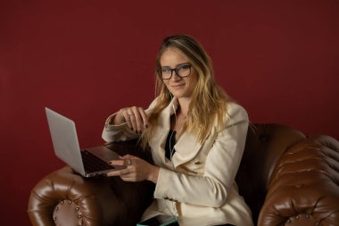 Evde dizüstü bilgisayar ve gazetelerle çalışan genç bir kadın klasik bir kahverengi koltuk ve kulaklıkla çalışıyor. Kırmızı boyalı duvar arka planı. Merkez ofis konsepti. Çalışmak için gri defter. Yüksek kalite fotoğraf