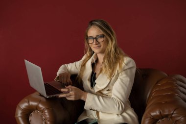 Evde dizüstü bilgisayar ve gazetelerle çalışan genç bir kadın klasik bir kahverengi koltuk ve kulaklıkla çalışıyor. Kırmızı boyalı duvar arka planı. Merkez ofis konsepti. Çalışmak için gri defter. Yüksek kalite fotoğraf