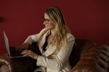 Evde dizüstü bilgisayar ve gazetelerle çalışan genç bir kadın klasik bir kahverengi koltuk ve kulaklıkla çalışıyor. Kırmızı boyalı duvar arka planı. Merkez ofis konsepti. Çalışmak için gri defter. Yüksek kalite fotoğraf