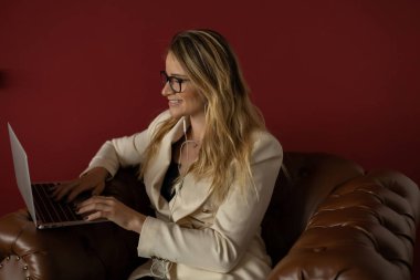 Evde dizüstü bilgisayar ve gazetelerle çalışan genç bir kadın klasik bir kahverengi koltuk ve kulaklıkla çalışıyor. Kırmızı boyalı duvar arka planı. Merkez ofis konsepti. Çalışmak için gri defter. Yüksek kalite fotoğraf