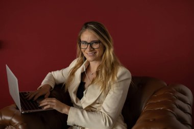 Evde dizüstü bilgisayar ve gazetelerle çalışan genç bir kadın klasik bir kahverengi koltuk ve kulaklıkla çalışıyor. Kırmızı boyalı duvar arka planı. Merkez ofis konsepti. Çalışmak için gri defter. Yüksek kalite fotoğraf