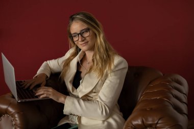 Evde dizüstü bilgisayar ve gazetelerle çalışan genç bir kadın klasik bir kahverengi koltuk ve kulaklıkla çalışıyor. Kırmızı boyalı duvar arka planı. Merkez ofis konsepti. Çalışmak için gri defter. Yüksek kalite fotoğraf