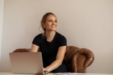 Evde dizüstü bilgisayarla çalışan genç bir kadın beyaz bir geçmişi olan kahverengi bir koltukta çalışıyor. Merkez ofis konsepti. Çalışmak için gri defter. Merkez ofis konsepti. Yüksek kalite fotoğraf