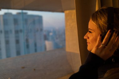 Kulağında kulaklıklarla müzik dinleyen genç bir kadının yan görüntüsü. Arka planda şehir manzarası var. Yüksek kalite fotoğraf. Cep telefonu, teknoloji, müzik, kentsel yaşam, iyi hisset.