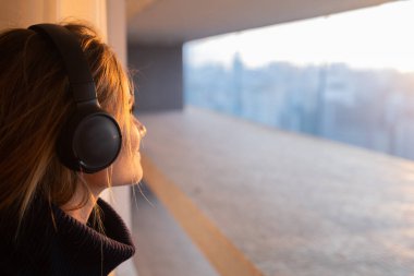 Kulağında kulaklıklarla müzik dinleyen genç bir kadının yan görüntüsü. Arka planda şehir manzarası var. Yüksek kalite fotoğraf. Cep telefonu, teknoloji, müzik, kentsel yaşam, iyi hisset.