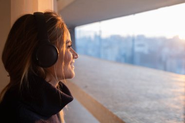 Kulağında kulaklıklarla müzik dinleyen genç bir kadının yan görüntüsü. Arka planda şehir manzarası var. Yüksek kalite fotoğraf. Cep telefonu, teknoloji, müzik, kentsel yaşam, iyi hisset.