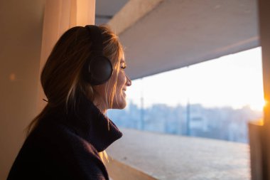 Kulağında kulaklıklarla müzik dinleyen genç bir kadının yan görüntüsü. Arka planda şehir manzarası var. Yüksek kalite fotoğraf. Cep telefonu, teknoloji, müzik, kentsel yaşam, iyi hisset.
