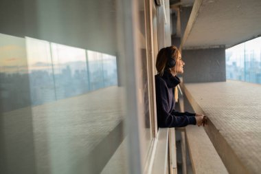 Kulağında kulaklıklarla müzik dinleyen genç bir kadının yan görüntüsü. Arka planda şehir manzarası var. Yüksek kalite fotoğraf. Cep telefonu, teknoloji, müzik, kentsel yaşam, iyi hisset.