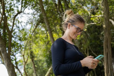 Gündüz vakti arka planda yeşil park olan akıllı telefon kullanan genç bir kadın. Yüksek kalite fotoğraf. Cep telefonu, teknoloji, şehir konsepti. Yüksek kalite fotoğraf