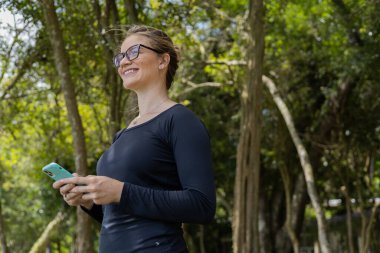 Gündüz vakti arka planda yeşil park olan akıllı telefon kullanan genç bir kadın. Yüksek kalite fotoğraf. Cep telefonu, teknoloji, şehir konsepti. Yüksek kalite fotoğraf