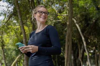 Gündüz vakti arka planda yeşil park olan akıllı telefon kullanan genç bir kadın. Yüksek kalite fotoğraf. Cep telefonu, teknoloji, şehir konsepti. Yüksek kalite fotoğraf