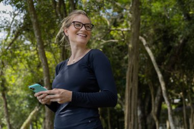 Gündüz vakti arka planda yeşil park olan akıllı telefon kullanan genç bir kadın. Yüksek kalite fotoğraf. Cep telefonu, teknoloji, şehir konsepti. Yüksek kalite fotoğraf