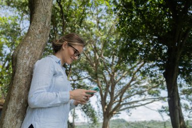 Gündüz vakti arka planda yeşil park olan akıllı telefon kullanan genç bir kadın. Yüksek kalite fotoğraf. Cep telefonu, teknoloji, şehir konsepti. Yüksek kalite fotoğraf