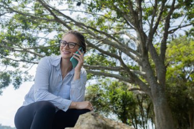 Gündüz vakti arka planda yeşil park olan akıllı telefon kullanan genç bir kadın. Yüksek kalite fotoğraf. Cep telefonu, teknoloji, şehir konsepti. Yüksek kalite fotoğraf