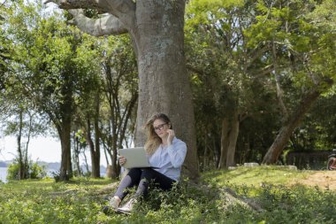 Gündüz vakti arka planda yeşil park olan bir dizüstü bilgisayar kullanan genç bir kadın. Yüksek kalite fotoğraf. Cep telefonu, teknoloji, şehir kavramı. 