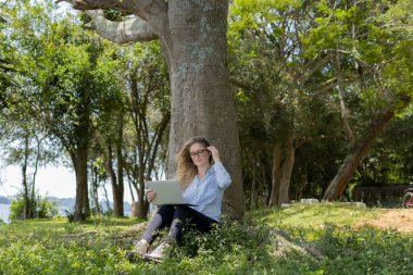 Gündüz vakti arka planda yeşil park olan bir dizüstü bilgisayar kullanan genç bir kadın. Yüksek kalite fotoğraf. Cep telefonu, teknoloji, şehir kavramı. 
