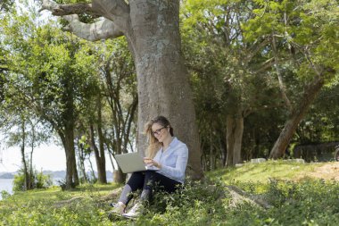 Gündüz vakti arka planda yeşil park olan bir dizüstü bilgisayar kullanan genç bir kadın. Yüksek kalite fotoğraf. Cep telefonu, teknoloji, şehir kavramı. 
