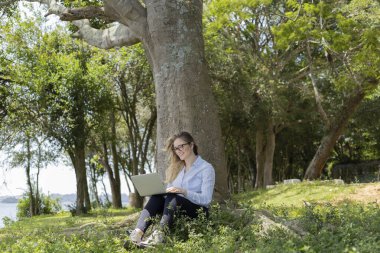 Gündüz vakti arka planda yeşil park olan bir dizüstü bilgisayar kullanan genç bir kadın. Yüksek kalite fotoğraf. Cep telefonu, teknoloji, şehir kavramı. 
