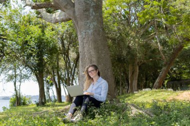 Gündüz vakti arka planda yeşil park olan bir dizüstü bilgisayar kullanan genç bir kadın. Yüksek kalite fotoğraf. Cep telefonu, teknoloji, şehir kavramı. 