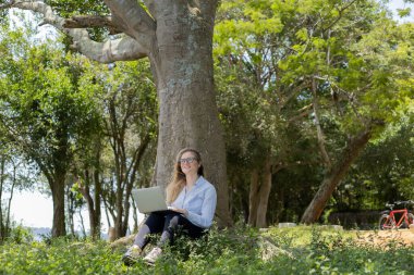 Gündüz vakti arka planda yeşil park olan bir dizüstü bilgisayar kullanan genç bir kadın. Yüksek kalite fotoğraf. Cep telefonu, teknoloji, şehir kavramı. 