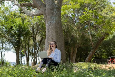Gündüz vakti arka planda yeşil park olan bir dizüstü bilgisayar kullanan genç bir kadın. Yüksek kalite fotoğraf. Cep telefonu, teknoloji, şehir kavramı. 