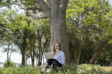 Gündüz vakti arka planda yeşil park olan bir dizüstü bilgisayar kullanan genç bir kadın. Yüksek kalite fotoğraf. Cep telefonu, teknoloji, şehir kavramı. 