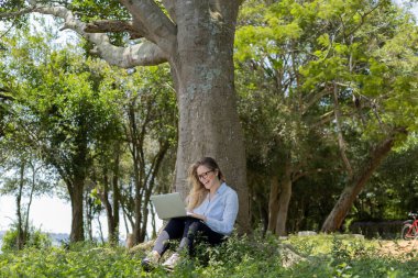 Gündüz vakti arka planda yeşil park olan bir dizüstü bilgisayar kullanan genç bir kadın. Yüksek kalite fotoğraf. Cep telefonu, teknoloji, şehir kavramı. 