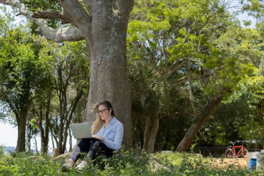 Gündüz vakti arka planda yeşil park olan bir dizüstü bilgisayar kullanan genç bir kadın. Yüksek kalite fotoğraf. Cep telefonu, teknoloji, şehir kavramı. 