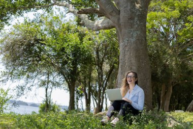 Gündüz vakti arka planda yeşil park olan bir dizüstü bilgisayar kullanan genç bir kadın. Yüksek kalite fotoğraf. Cep telefonu, teknoloji, şehir kavramı. 