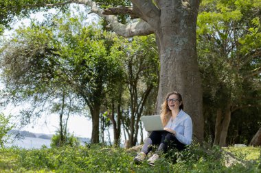 Gündüz vakti arka planda yeşil park olan bir dizüstü bilgisayar kullanan genç bir kadın. Yüksek kalite fotoğraf. Cep telefonu, teknoloji, şehir kavramı. 