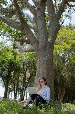 Gündüz vakti arka planda yeşil park olan bir dizüstü bilgisayar kullanan genç bir kadın. Yüksek kalite fotoğraf. Cep telefonu, teknoloji, şehir kavramı. 