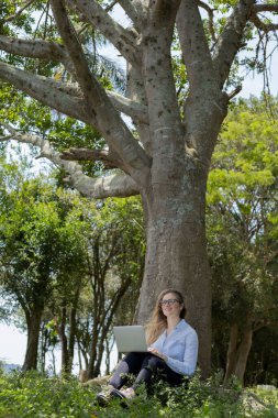 Gündüz vakti arka planda yeşil park olan bir dizüstü bilgisayar kullanan genç bir kadın. Yüksek kalite fotoğraf. Cep telefonu, teknoloji, şehir kavramı. 