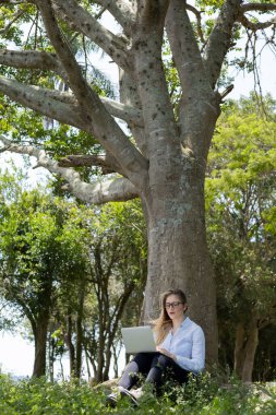 Gündüz vakti arka planda yeşil park olan bir dizüstü bilgisayar kullanan genç bir kadın. Yüksek kalite fotoğraf. Cep telefonu, teknoloji, şehir kavramı. 