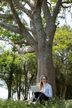 Gündüz vakti arka planda yeşil park olan bir dizüstü bilgisayar kullanan genç bir kadın. Yüksek kalite fotoğraf. Cep telefonu, teknoloji, şehir kavramı. 
