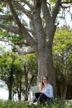 Gündüz vakti arka planda yeşil park olan bir dizüstü bilgisayar kullanan genç bir kadın. Yüksek kalite fotoğraf. Cep telefonu, teknoloji, şehir kavramı. 