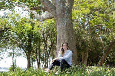 Gündüz vakti arka planda yeşil park olan akıllı telefon kullanan genç bir kadın. Yüksek kalite fotoğraf. Cep telefonu, teknoloji, şehir konsepti. Yüksek kalite fotoğraf