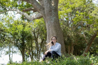 Gündüz vakti arka planda yeşil park olan akıllı telefon kullanan genç bir kadın. Yüksek kalite fotoğraf. Cep telefonu, teknoloji, şehir konsepti. Yüksek kalite fotoğraf