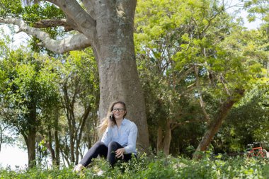 Gündüz vakti arka planda yeşil park olan akıllı telefon kullanan genç bir kadın. Yüksek kalite fotoğraf. Cep telefonu, teknoloji, şehir konsepti. Yüksek kalite fotoğraf