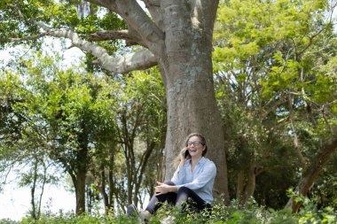 Gündüz vakti arka planda yeşil park olan akıllı telefon kullanan genç bir kadın. Yüksek kalite fotoğraf. Cep telefonu, teknoloji, şehir konsepti. Yüksek kalite fotoğraf