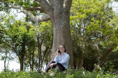 Gündüz vakti arka planda yeşil park olan akıllı telefon kullanan genç bir kadın. Yüksek kalite fotoğraf. Cep telefonu, teknoloji, şehir konsepti. Yüksek kalite fotoğraf