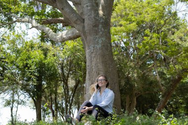 Gündüz vakti arka planda yeşil park olan akıllı telefon kullanan genç bir kadın. Yüksek kalite fotoğraf. Cep telefonu, teknoloji, şehir konsepti. Yüksek kalite fotoğraf