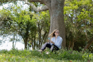 Gündüz vakti arka planda yeşil park olan akıllı telefon kullanan genç bir kadın. Yüksek kalite fotoğraf. Cep telefonu, teknoloji, şehir konsepti. Yüksek kalite fotoğraf
