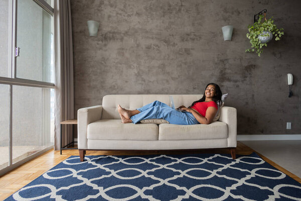 Young black woman working at home with laptop on her lap lying down on her couch in the living room.  Home office concept. Gray notebook for working. High quality photo
