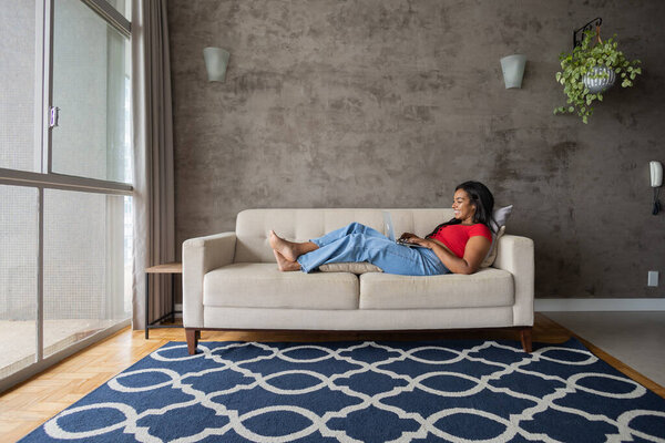 Young black woman working at home with laptop on her lap lying down on her couch in the living room.  Home office concept. Gray notebook for working. High quality photo