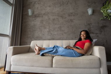 Genç siyahi kadın evde dizüstü bilgisayarıyla çalışıyor oturma odasında kanepede uzanıyor. Merkez ofis konsepti. Çalışmak için gri defter. Yüksek kalite fotoğraf