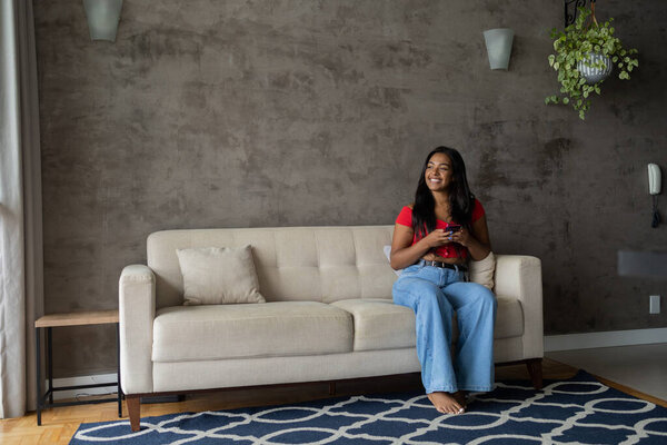 Young black woman working at home with smartphone sitting on her couch in the living room.  Home office concept. High quality photo