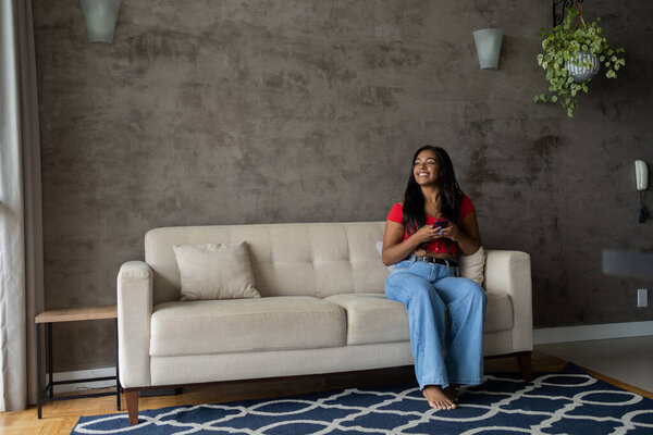 Young black woman working at home with smartphone sitting on her couch in the living room.  Home office concept. High quality photo